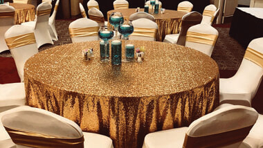 Round table with gold tablecloth, teal votive, and cream chair covers.