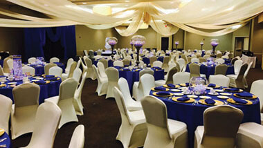 White material is draped from the ceiling over circular tables covered in dark blue table cloths and surrounded by chairs covered in white.
