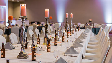 A family-style reception setup featuring long tables in white linen with tall candle sticks down the center.